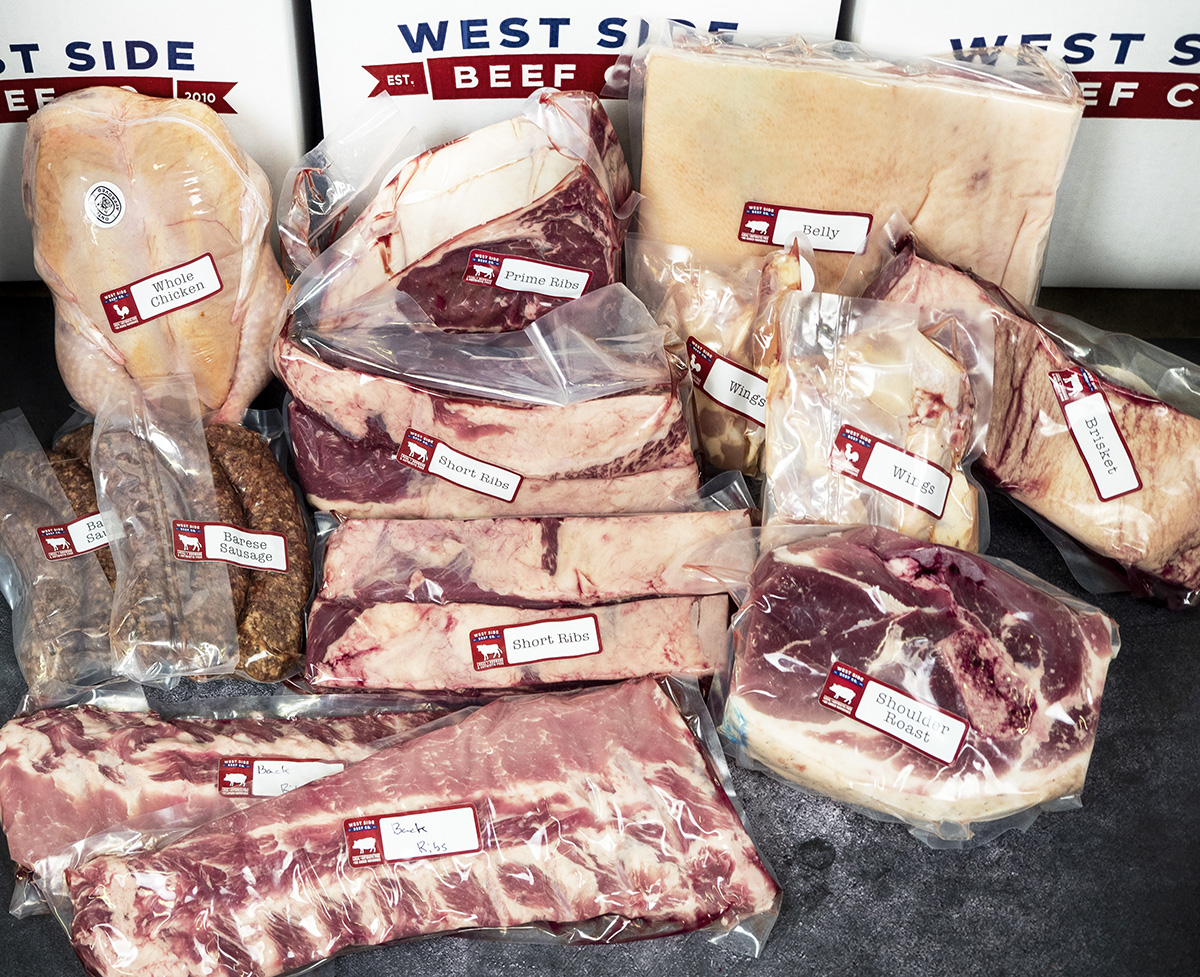 Assorted packaged meats, including whole chicken, ribs, pork belly, shoulder roast, sausages, and wings, arranged in branded boxes.