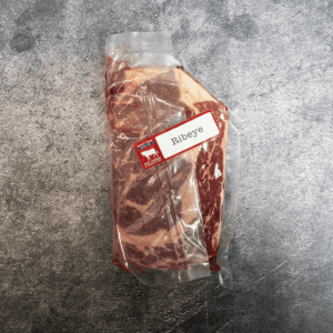 Vacuum-sealed ribeye steak with visible marbling, labeled and placed on a gray surface.
