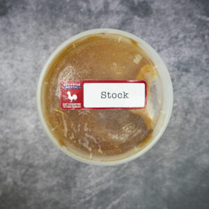 Round plastic container of frozen chicken stock with a "Stock" label on a gray textured surface.