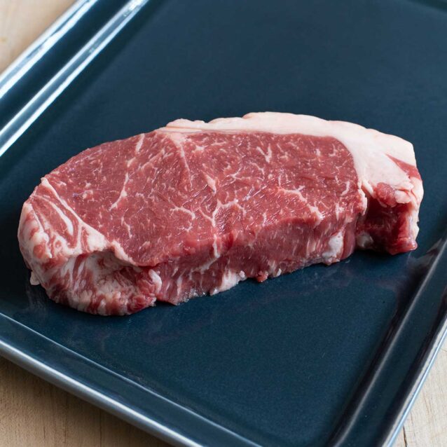 Thick-cut striploin steak with visible marbling and fat cap, placed on a dark plate.