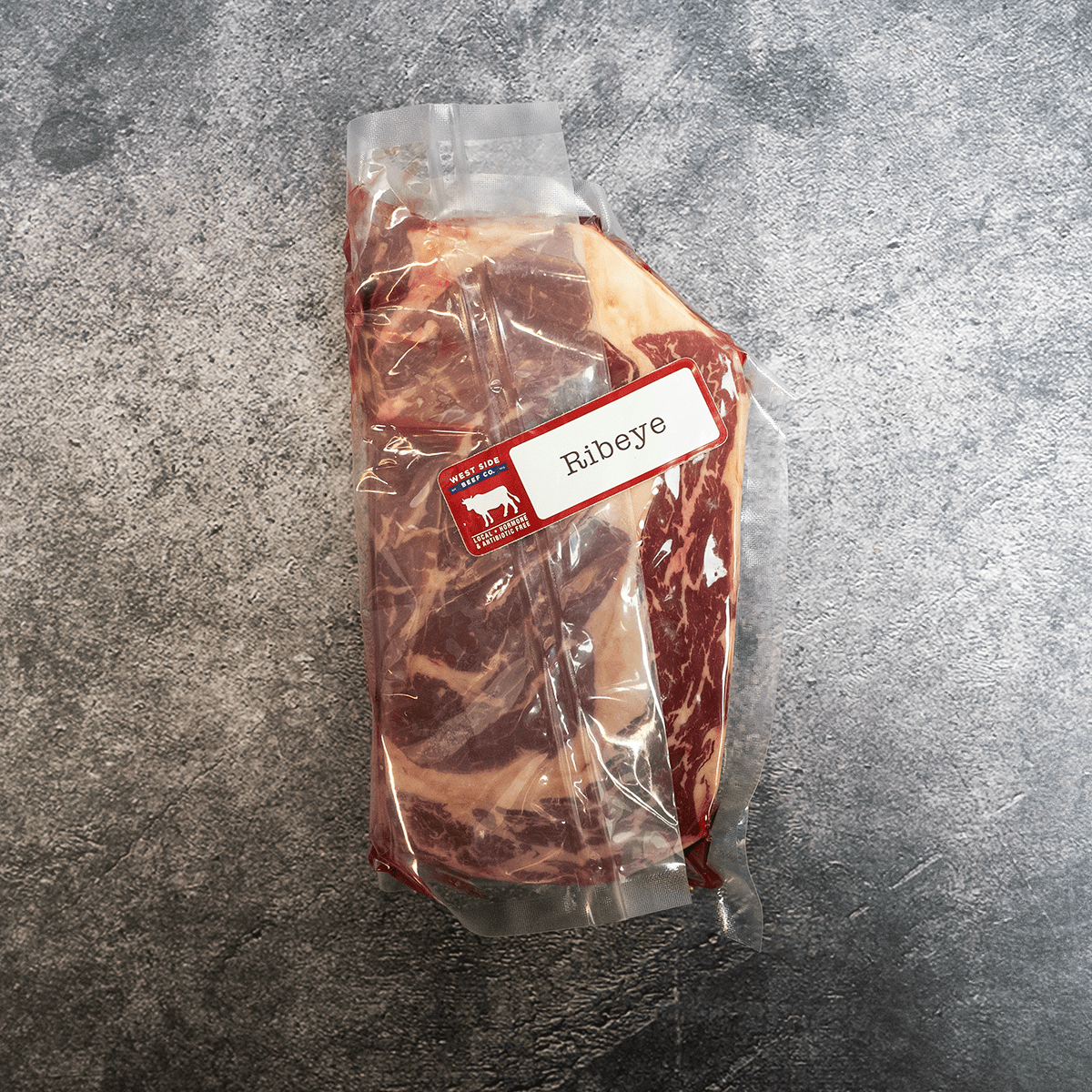 Vacuum-sealed ribeye steak with visible marbling, labeled and placed on a gray surface.