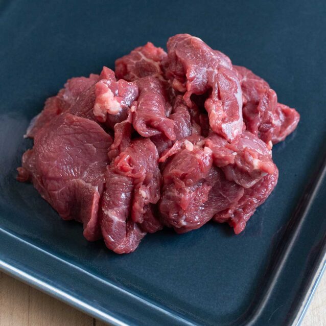 Raw beef stir-fry strips piled on a dark tray, showing thin cuts with light marbling.