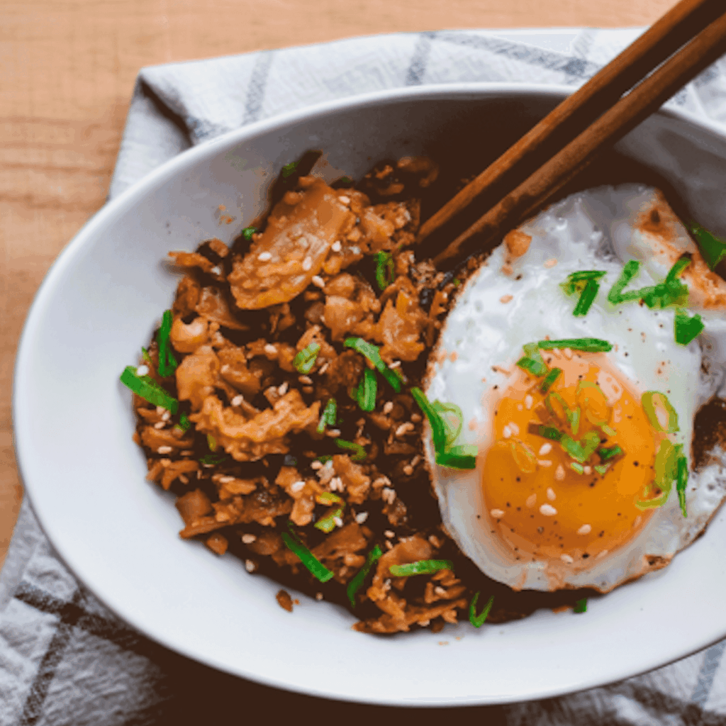 Bowl of fried rice with bacon, kimchi, and a sunny-side-up egg, garnished with green onions.