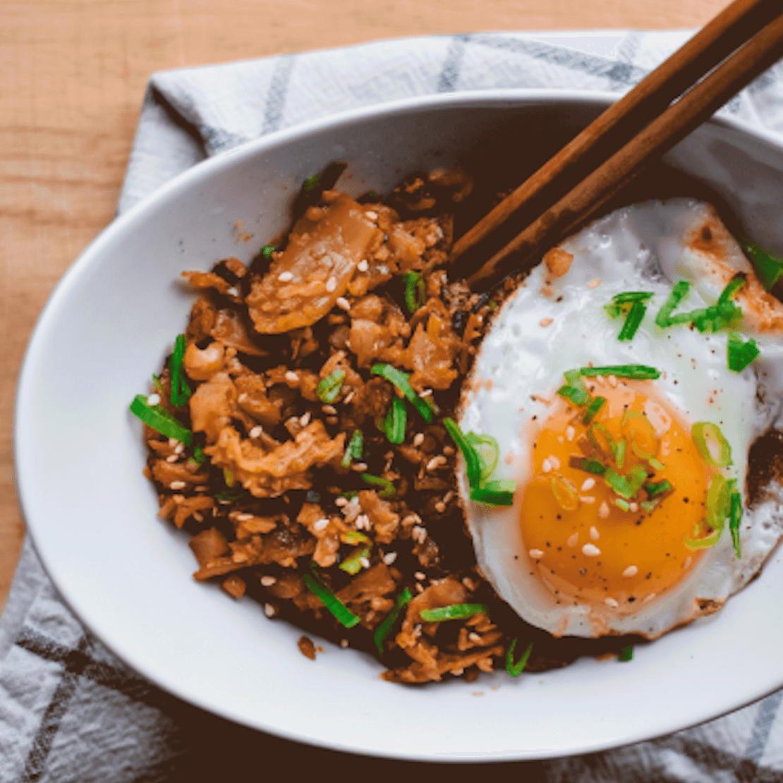 Bowl of fried rice with bacon, kimchi, and a sunny-side-up egg, garnished with green onions.
