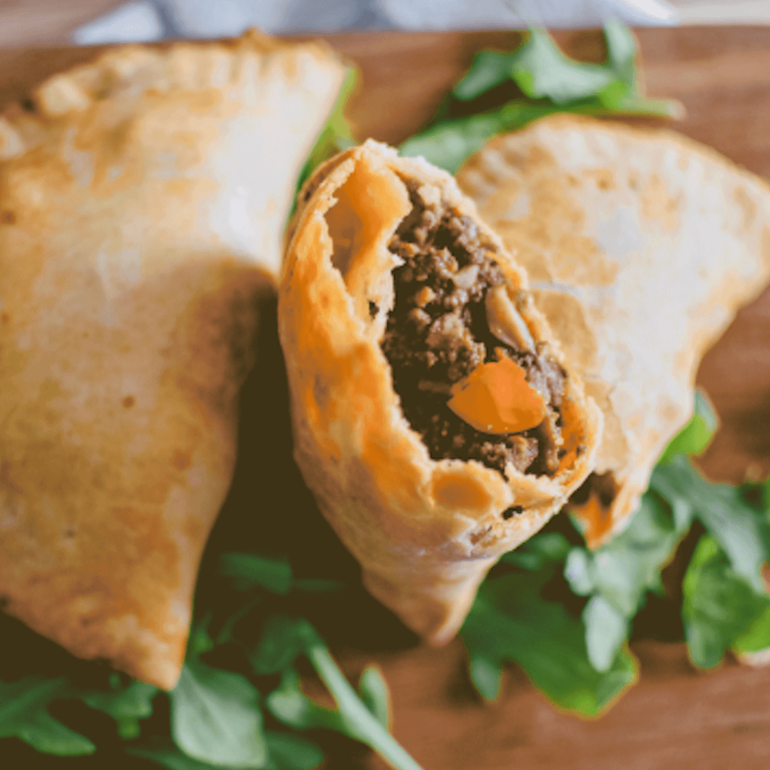 Golden-brown hand pies with a visible beef filling, served on a wooden surface with greens.